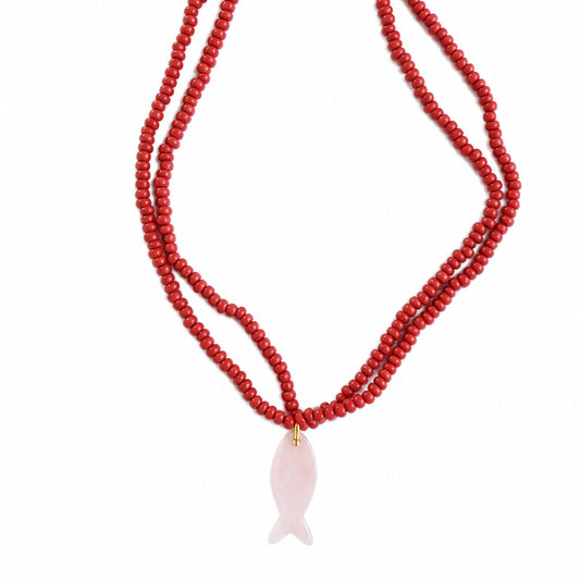 A double-strand red beaded necklace with a pale pink fish-shaped pendant, laid flat on a textured concrete surface.