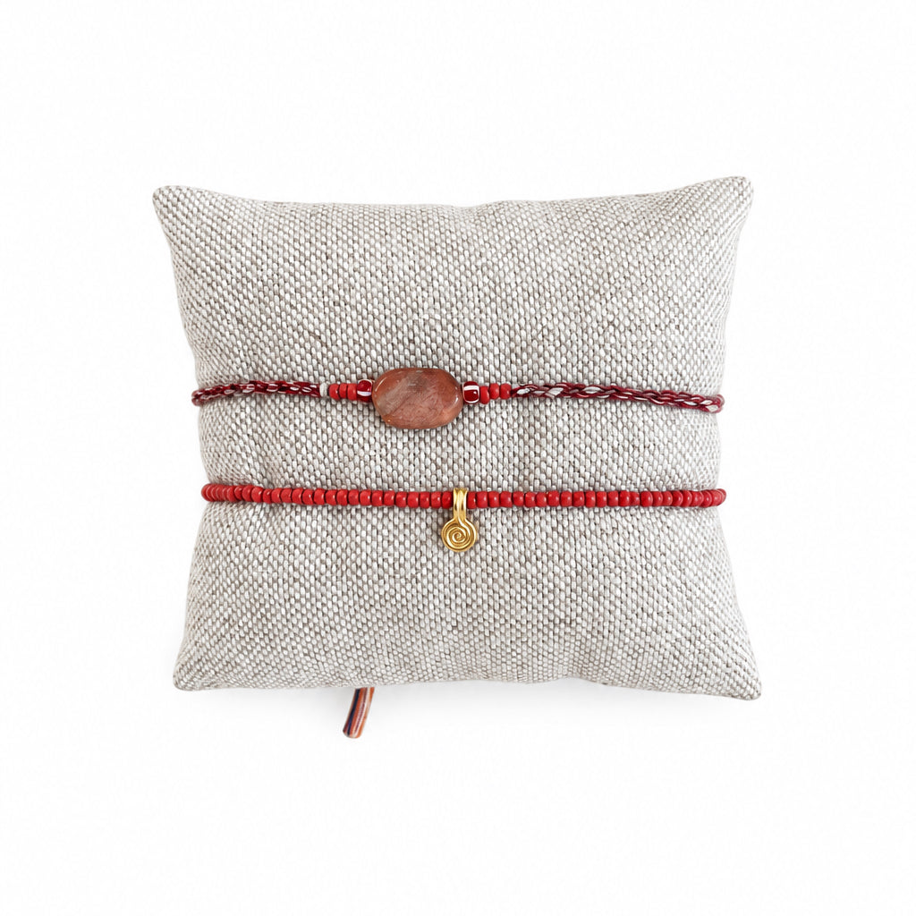 Two red cord bracelets, one with a small brown wooden bead and the other with a tiny gold charm, displayed on a beige woven pillow over a concrete surface.