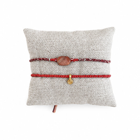 Two red cord bracelets, one with a small brown wooden bead and the other with a tiny gold charm, displayed on a beige woven pillow over a concrete surface.